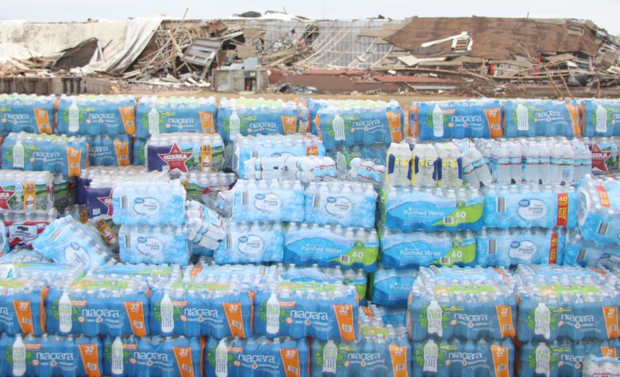 A wall of cases of water bottles lay in the foreground as a damaged building sits in the background in Rolling Fork, Mississippi on March 31, 2023.